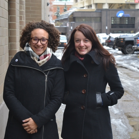 Barbara Hilden and Rebecca Jade standing side by side in an alley, both in long black coats